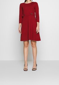 Czerwona sukienka z dopasowanym gorsetem, rękawami do łokcia i rozkloszowaną spódnicą. Posiada metalowe akcenty w talii oraz gładką fakturę materiału.