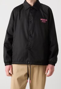 Czarna kurtka przeciwwiatrowa z kołnierzem, zatrzaskami, elastycznymi mankietami i regulowanym dołem. Posiada różowe logo na piersi.