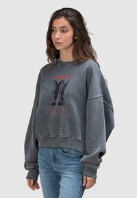 Bluza z szarego węgla o luźnym kroju, z nadrukiem graficznym i prążkowanymi wykończeniami. Połączona z jasnoniebieskimi jeansami z przetarciami.