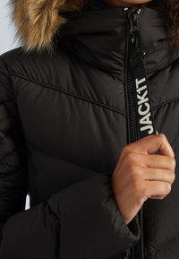 Schwarze Steppjacke mit Reißverschluss vorne, ausgestattet mit einem kunstpelzbesetzten Kapuzenrand und diagonaler Steppmusterung. Logoband entlang des Reißverschlusses.