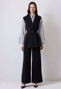Black tailored vest with a belted waist over a gray shirt, featuring wide sleeves. Paired with black wide-leg trousers and pointed heels.