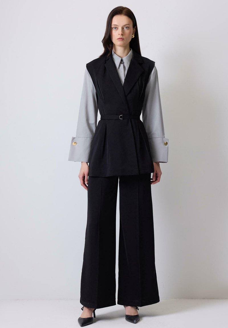 Black tailored vest with a belted waist over a gray shirt, featuring wide sleeves. Paired with black wide-leg trousers and pointed heels.