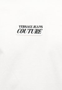 Λευκό ύφασμα με ορθογώνιο μπαλώμα που εμφανίζει τη φράση "VERSACE JEANS COUTURE" με κεφαλαία μαύρα γράμματα.