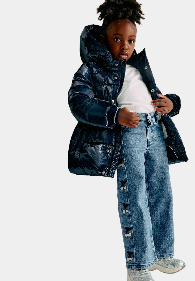 Marinblå puffjacka med hög krage, i kombination med ljusblå vida jeans med blommiga sidoknappar och grå sneakers.