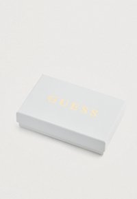 Scatola rettangolare bianca con superficie texture, con la parola "GUESS" in lettere dorate sulla parte superiore.