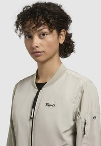 Jachetă bomber bej cu guler ribbed, fermoar frontal și logo brodat. Include un buzunar cu fermoar pe mâneca stângă. Material neted, ușor.