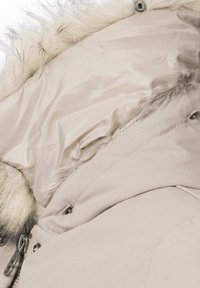 Beige parka with a faux fur-lined hood, featuring smooth fabric, silver hardware, and a quilted inner lining. Zipper and snaps visible.