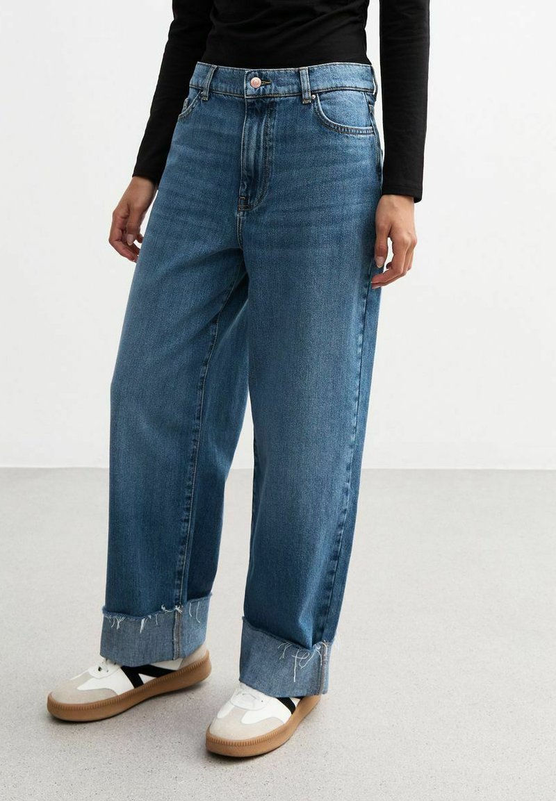 Vaqueros de mezclilla de cintura alta, de pierna ancha y color azul claro, con dobladillos desgastados, cinco bolsillos y combinados con zapatillas blancas y beige.
