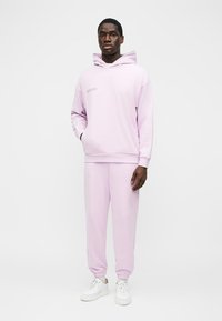 Mężczyzna stojący w jasnofioletowej bluzie z kapturem i dopasowanych spodniach dresowych oraz białych tenisówkach na jednolitym białym tle.