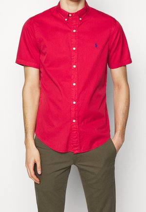 Homme portant une chemise à manches courtes boutonnée rouge vif et un pantalon vert olive, une main dans la poche, sur un fond blanc.
