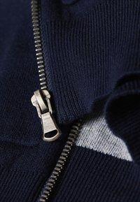 Marine gebreid jack met een rits aan de voorkant, voorzien van een zilveren metalen rits en een geribbelde textuur. Accentstukken van grijze stof zijn zichtbaar aan de zijkanten.