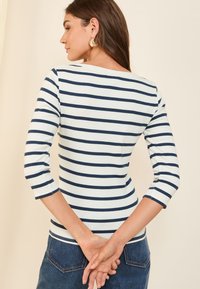 Gestreiftes Langarmshirt mit Marineblauen Streifen auf weißem Grund, ausgestattet mit einem Bootsausschnitt und einer taillierten Passform, getragen mit Jeans.