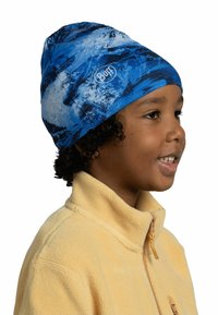 Blue and black patterned beanie made from stretchy fabric, fitted at the base, featuring a circular logo on the side. Child wearing a yellow fleece jacket.