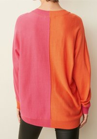 Suéter bicolor que presenta un lado izquierdo rosa y un lado derecho naranja, hecho de un material de punto suave, con un ajuste holgado y puños y dobladillo acanalados.