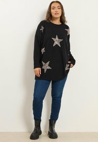 Un pull noir avec des appliqués d'étoiles en sequins aux tons argentés, à coupe décontractée, manches longues et poches latérales. Porté avec un jean bleu et des bottes noires.