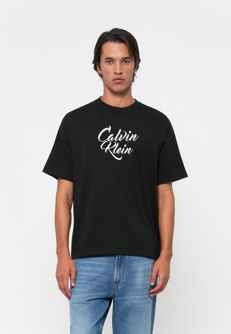 Mees, kes kannab musta lühikeste varrukatega T-särki, millel on valge "Calvin Klein" kiri, ja heledaid siniseid teksaseid, seisab ühtlase valge taustaga.