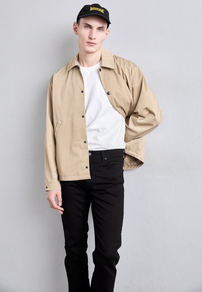 Beige Jacke mit schwarzen Knöpfen, weißem Hemd und schwarzen Jeans. Das Model trägt eine schwarze Mütze mit gelbem Text und posiert vor einem neutralen Hintergrund.