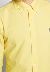 Żółta koszula na guziki z teksturowanym wykończeniem, posiadająca kołnierzyk oraz małe, niebieskie, haftowane logo. Widocznych jest pięć białych guzików.