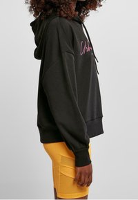 Černá mikina s kapucí, na přední straně s růžovým grafickým textem, kombinovaná s žlutými cyklistickými šortkami a volně střiženým stylem.