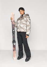 Spīdīga sudrabota pūdera jaka ar dimanta rakstu, melnas slēpošanas bikses un melni slēpošanas zābaki. Rokās tur rozā un melnas krāsas slēpes ar stiprinājumiem. Uz galvas slēpošanas brilles.