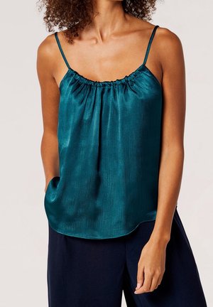 Camisole en satin bleu sarcelle avec fines bretelles réglables, encolure froncée et silhouette fluide, associée à un pantalon large foncé.