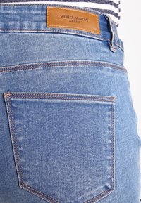 Ljusblå denimjeans med en brun läderlapp märkt "VERO MODA." Bakfickan har standardstickning och en något sliten textur.