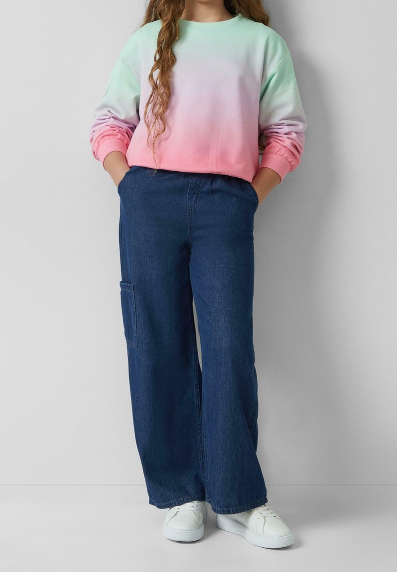 Enfant portant un sweat-shirt ombré pastel, un jean large en denim foncé avec une poche latérale et des baskets blanches, debout contre un mur uni.
