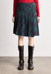 Πράσινη καρό φούστα με πτυχώσεις και λευκή δαντελένια λεπτομέρεια στο τελείωμα, συνδυασμένη με μαύρες δερμάτινες μπότες ως το γόνατο που διαθέτουν αξεσουάρ με λουριά.