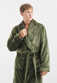 Robe in fleece verde oliva, morbido e peloso, con colletto a scialle, chiusura con nastro davanti e due tasche a toppa. Texture soffice e maniche lunghe.