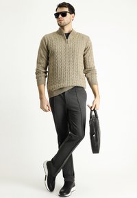 Beige Kabelstrickpullover mit hohem Kragen, dunkelgraue schmal geschnittene Hose, schwarze Sneakers und eine schwarze Lederaktentasche.