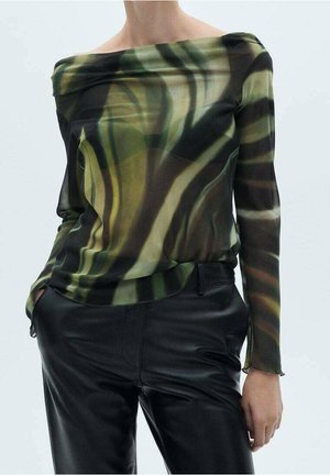 Persona con un top de hombros descubiertos, de manga larga, con un patrón abstracto verde y negro, y pantalones de cuero negros, de pie con una mano en el bolsillo.