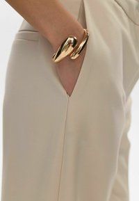 Bracciale a cuff in metallo dorato con un design liscio e curvo, indossato su una mano parzialmente visibile in una tasca di pantaloni beige.