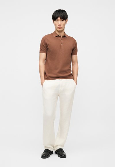 Un homme se tient les mains dans les poches, portant un polo marron à manches courtes, un pantalon blanc et des chaussures noires, sur un fond blanc uni.