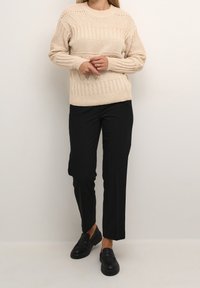Beige gestrickter Pullover mit strukturierten Mustern, kombiniert mit schwarzen maßgeschneiderten Hosen und klobigen schwarzen Mokkasins.