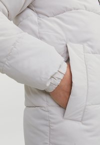 Lätt grå pufferjacka med elastisk manschett och en sidoficka. Materialet verkar slätt med kviltad isoleringsdetalj.