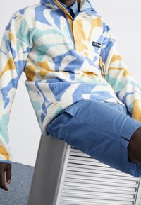 Pull-over en polaire blanc avec des motifs abstraits bleu, jaune et orange. Doté d'une fermeture éclair sur le devant et d'une poche sur la poitrine. Porté avec un short bleu.