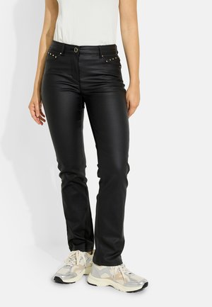 Mujer que lleva pantalones negros de cuero sintético con tachuelas doradas en los bolsillos y zapatillas blancas con detalles en gris y beige.