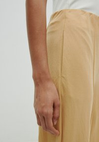Lichtbeige, hoge taille broeken met een gladde textuur en zijplooien. Een hand rust op de stof, waardoor het ontwerp en de kleur worden benadrukt.