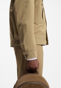 Gaiši brūna jaka ar gludu tekstūru, kas aprīkota ar sānu kabatu, pogām aizdares aprocēm un personu, kas tur bēša krāsas mugursomu ar rāvējslēdzēju.