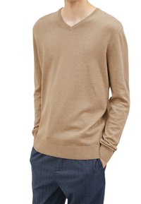 Uomo che indossa un maglione a V di colore marrone chiaro e pantaloni blu scuro con le mani nelle tasche, in piedi contro uno sfondo bianco.