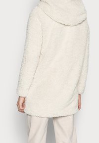 Person klädd i en långärmad, off-white, fluffig fleecejacka med bred krage, tillsammans med ljusa byxor, sedd bakifrån.