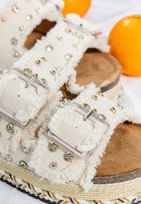 Plattform-Sandaletten mit cremefarbenen Stoffriemen, verziert mit klaren und goldenen Nieten, mit ausgefransten Kanten und einer Korksohle. Goldfarbener geflochtener Rand.