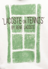 Sweatshirt blanc avec une grande impression verte ressemblant à un court de tennis, avec le texte "LACOSTE on TENNIS" par Rene Lacoste.