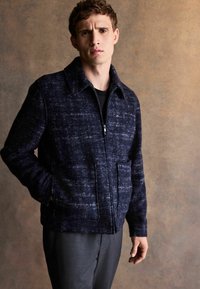 Marineblaue strukturierte Jacke mit Stehkragen, Frontreißverschluss und Seitentaschen, gefertigt aus einem Wollmischgewebe mit dezentem Muster.