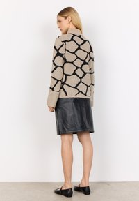 Beiger Pullover mit schwarzem geometrischem Muster und hohem Kragen. Kombiniert mit einem schwarzen, strukturierten Lederrock, der bis zur Mitte des Oberschenkels reicht.