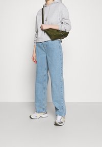 Felpa grigia corta, jeans azzurri larghi e sneakers bianche con accenti gialli. Borsa a spalla verde oliva con dettagli in argento.