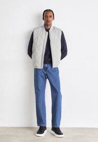 Gilet trapuntato grigio chiaro sopra un maglione blu navy, abbinato a jeans dritti blu e sneakers nere. Design semplice senza decorazioni.