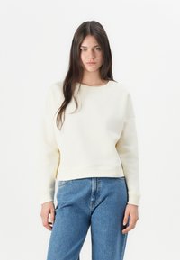 Cream-colored sweatshirt with a round neck, dropped shoulders, and ribbed hem, paired with light blue denim jeans. Minimalist design.