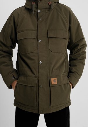 Olivengrøn hættejakke med fire lommer med klapper foran, knappelukning og lynlås, båret af person med hænderne i lommerne.