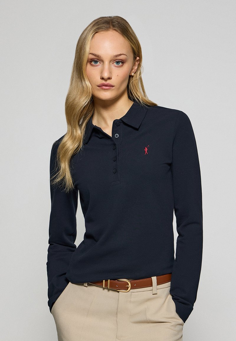 Långärmad polo i marinblå bomull, med en treknappsslutning och en liten röd logotyp. Brun bälte i kombination med beige byxor.